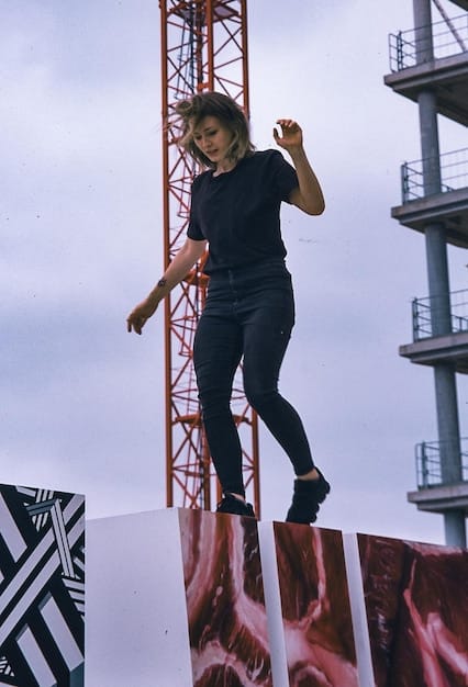 A behind-the-scenes shot of a complex action sequence being filmed. Stunt performers are suspended on wires, while the director and camera crew monitor the scene from below. The set is filled with smoke and debris, creating a dynamic and chaotic atmosphere.